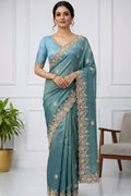Woman wearing a teal saree with gold embroidery in a minimalistic room.