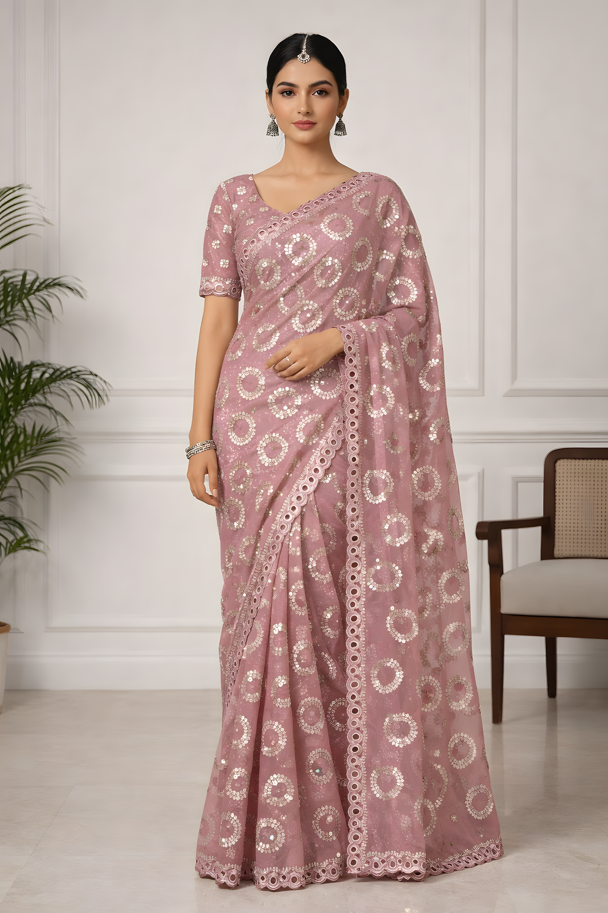Woman wearing a pink saree with white patterns in a room with a white wall and chair.