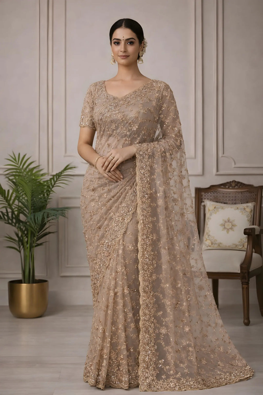 Woman wearing a beige saree with intricate patterns in a room with a plant and chair.