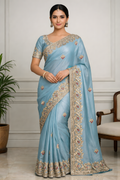 Woman wearing a light blue saree with intricate patterns in a room setting.