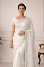 Woman wearing a white saree with a subtle border in a room with a plant and chair.